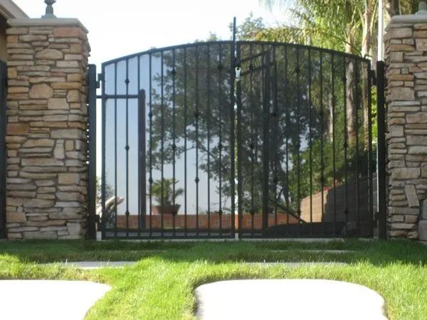 Black wrought-iron gate between stone pillars, reflecting trees and a building.