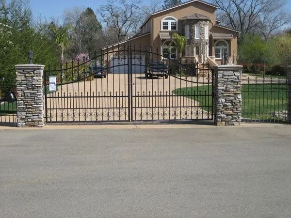 Wrought iron driveway gates with stone pillars leading to a house. A driveway and cars are visible.