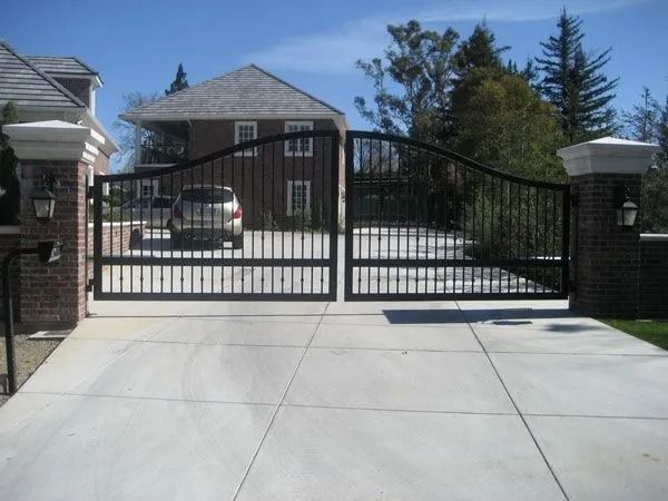 Black wrought-iron driveway gates with brick pillars, opening to a concrete drive, a house, and a parked car.