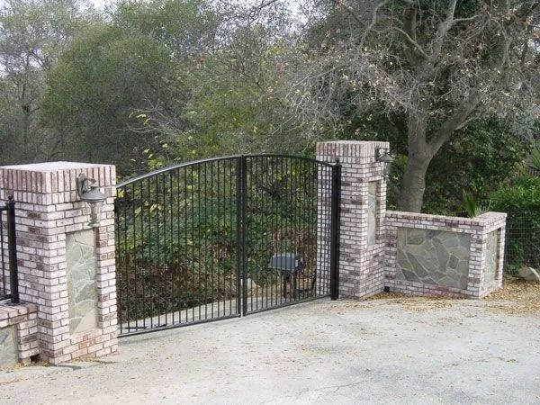 Black iron gate flanked by brick pillars with stone accents; driveway, trees in background.