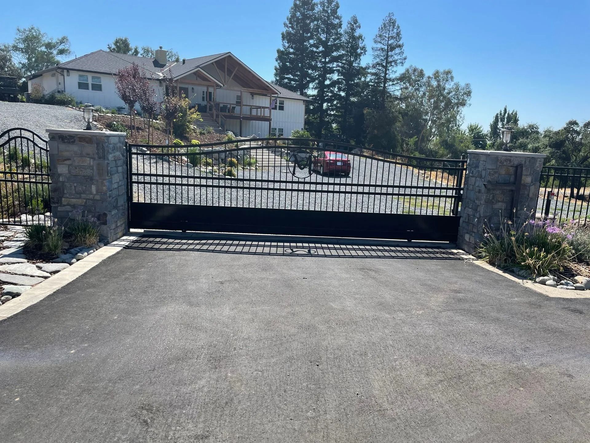 Black wrought-iron gate in front of a house, set between stone columns, on a paved driveway under a sunny sky.