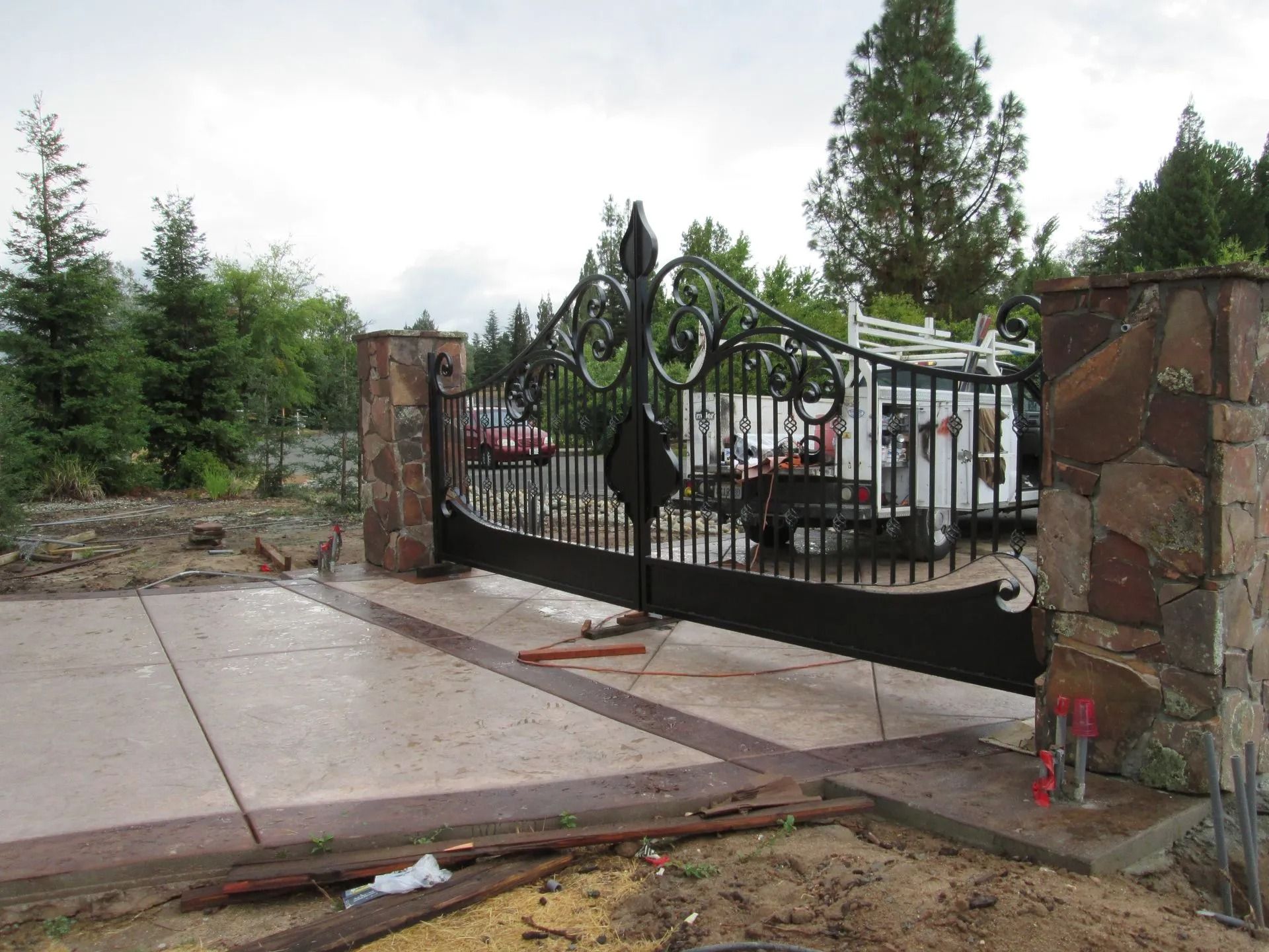 Ornate black metal driveway gate with stone pillars, set in concrete, outdoors.