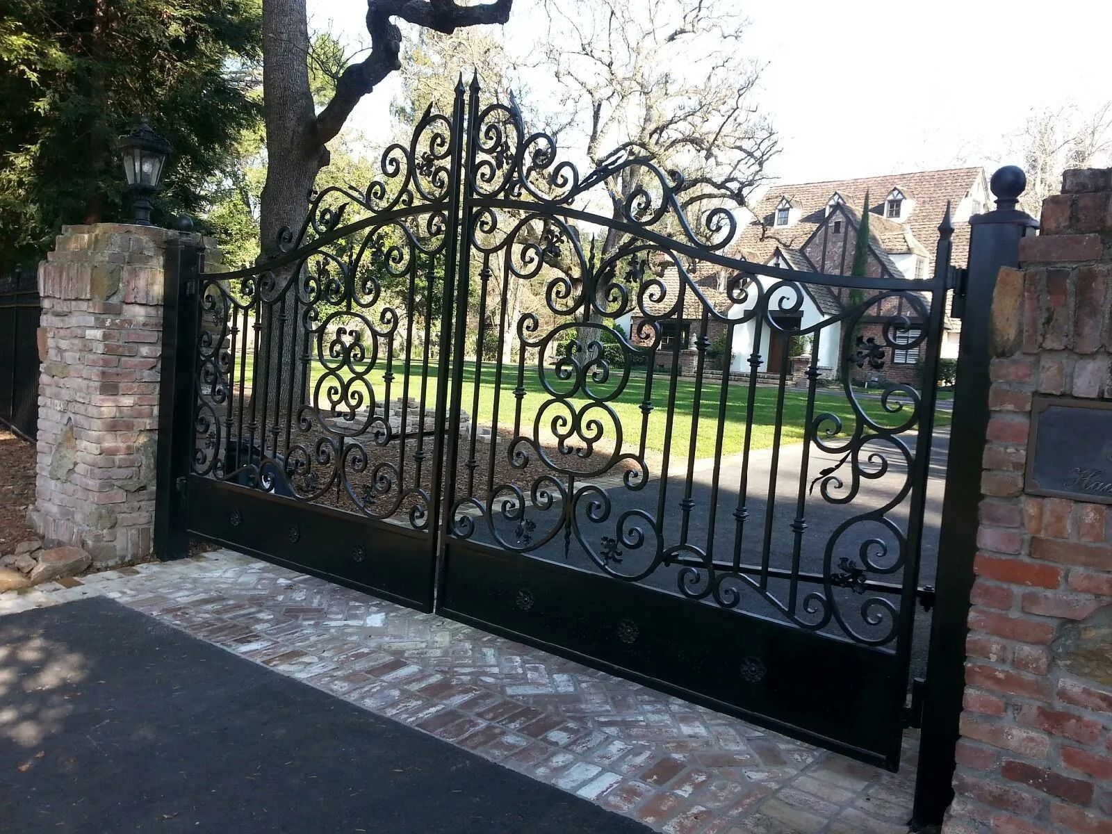 Black ornate iron gates on brick pillars, leading to a house. Paved driveway.