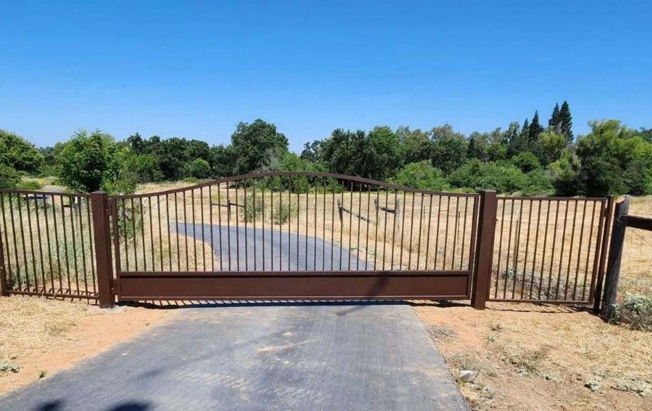 Brown metal gate across a paved driveway, in front of trees under a blue sky.