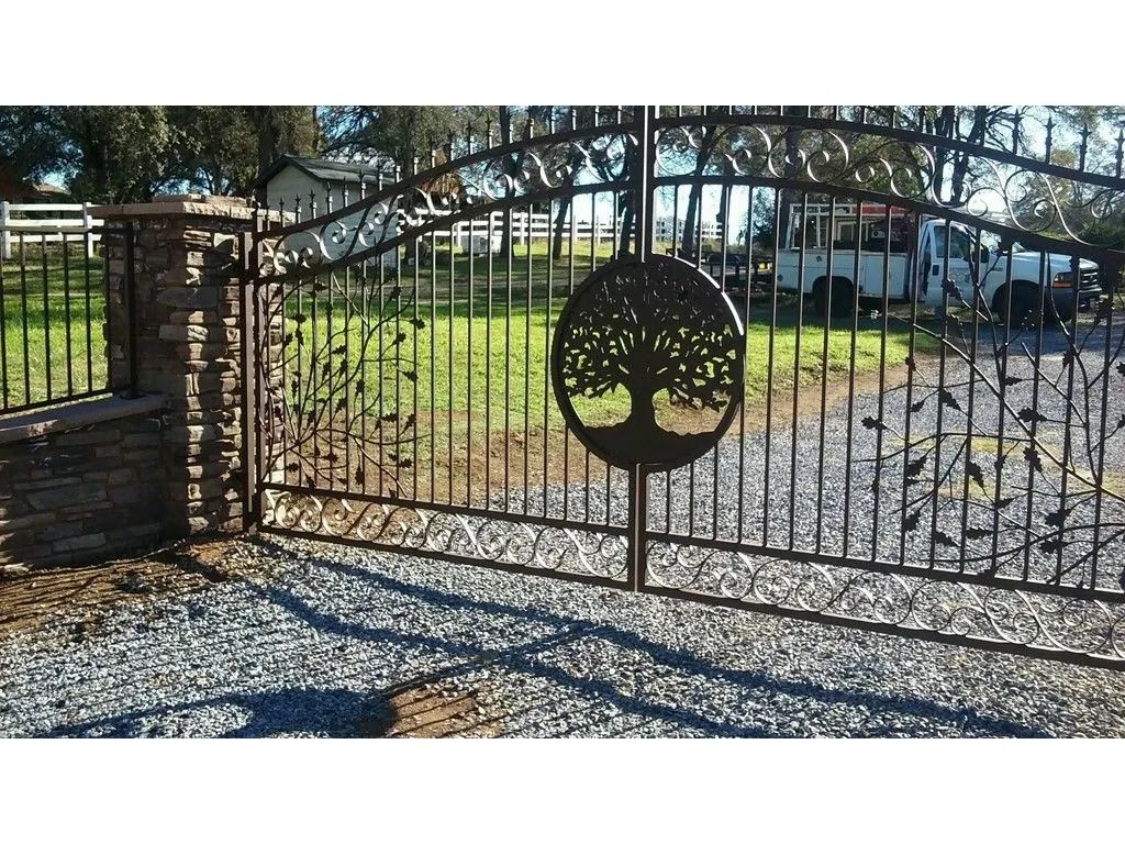 Black metal gate with tree design, set on gravel, leading to a grassy area with a white fence and a truck.