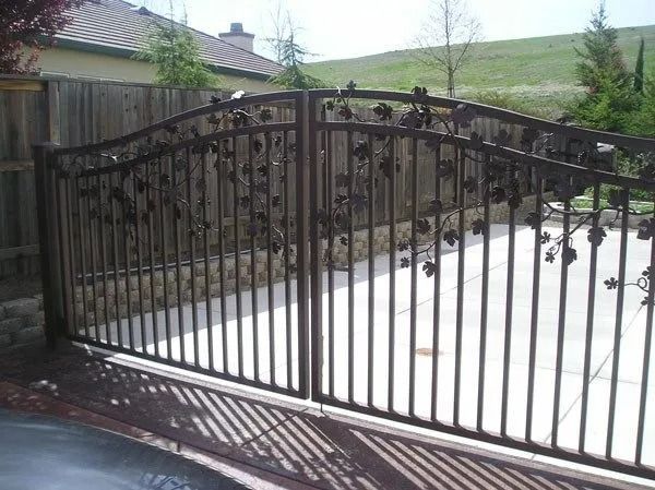 Dark metal gate with leaf designs, arched over a driveway, with a wooden fence and grassy hill in the background.
