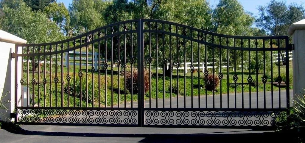 Black wrought iron gate with decorative details. Behind the gate is green foliage and a paved driveway.
