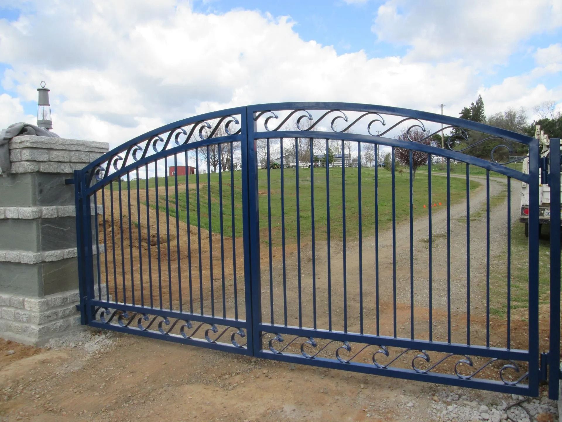 Blue wrought iron driveway gate on brick columns, leading to a grassy yard under a cloudy sky.