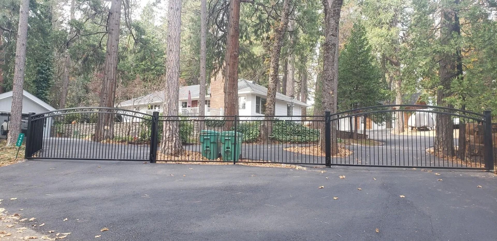 A house is set back behind a black iron fence and large trees. Green recycling bins are visible.