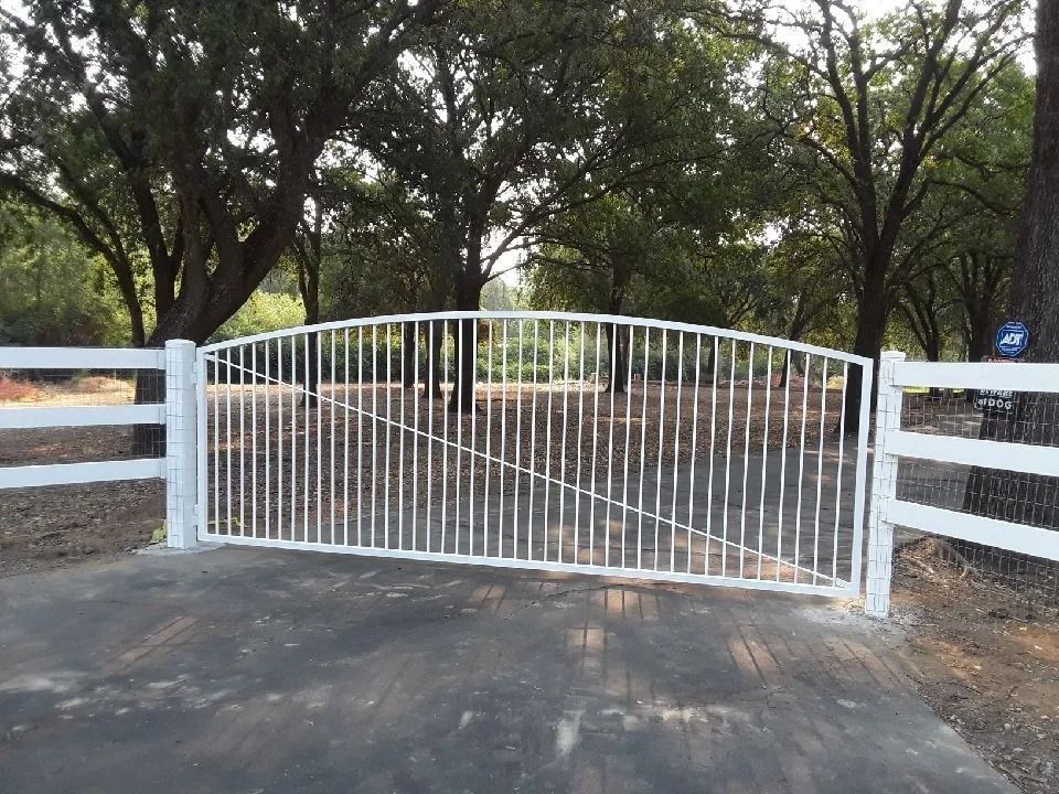 White metal gate with vertical bars, set between white fence posts, in front of trees.
