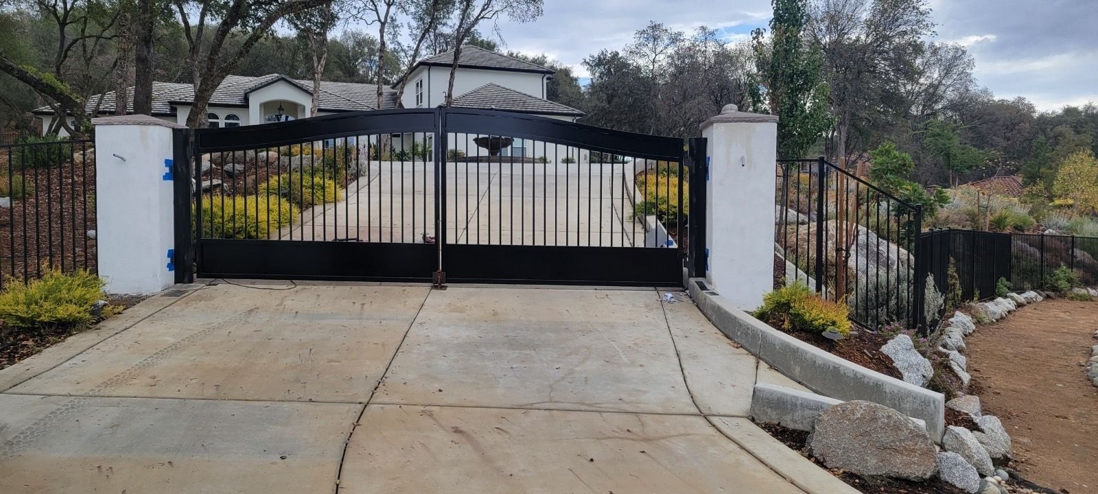 Black gate at the entrance of a house, set on a concrete driveway with a garden along the right.
