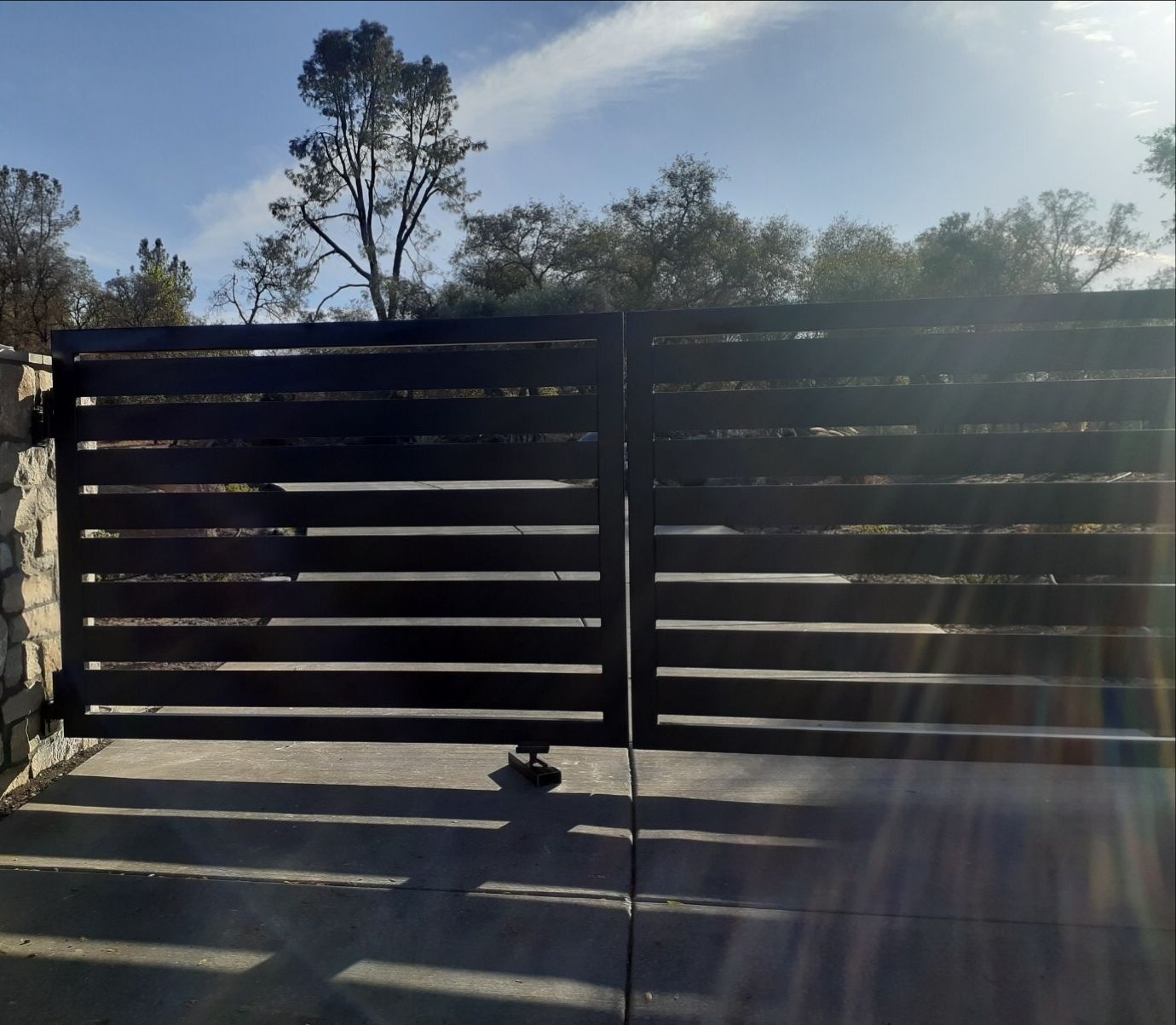 Black horizontal slat gates, set in stone, casting shadows. Trees and blue sky in background.