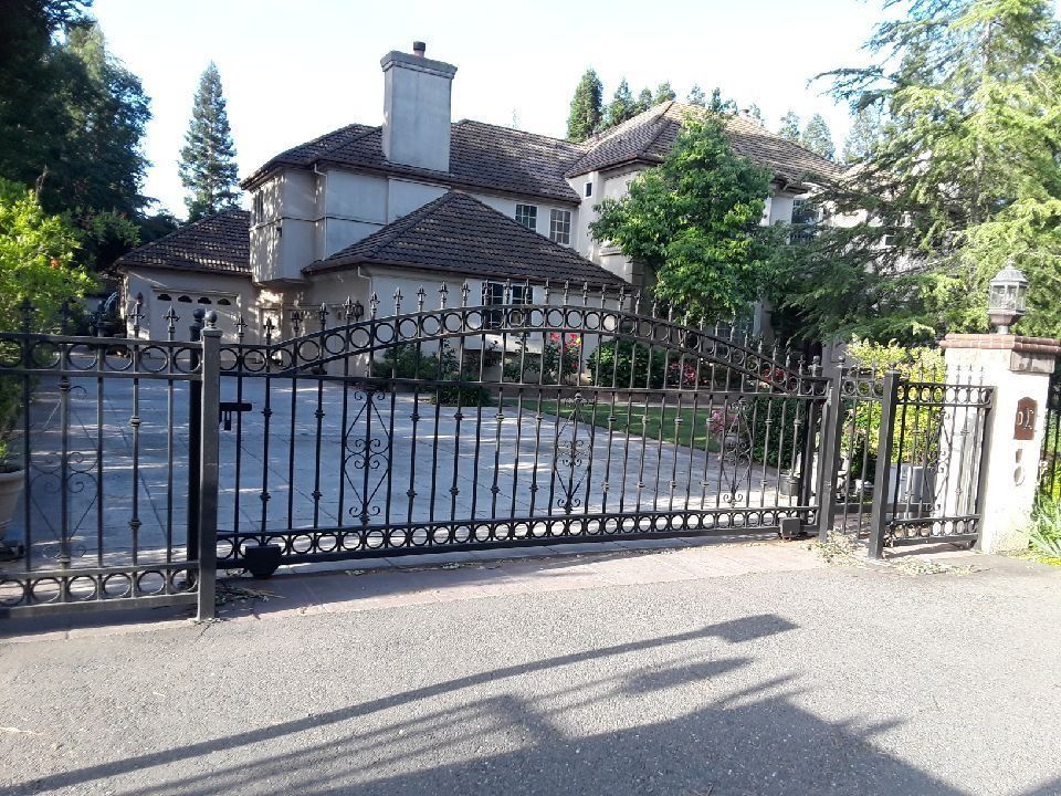 Black iron gate in front of a large, light-colored house with a tiled roof.