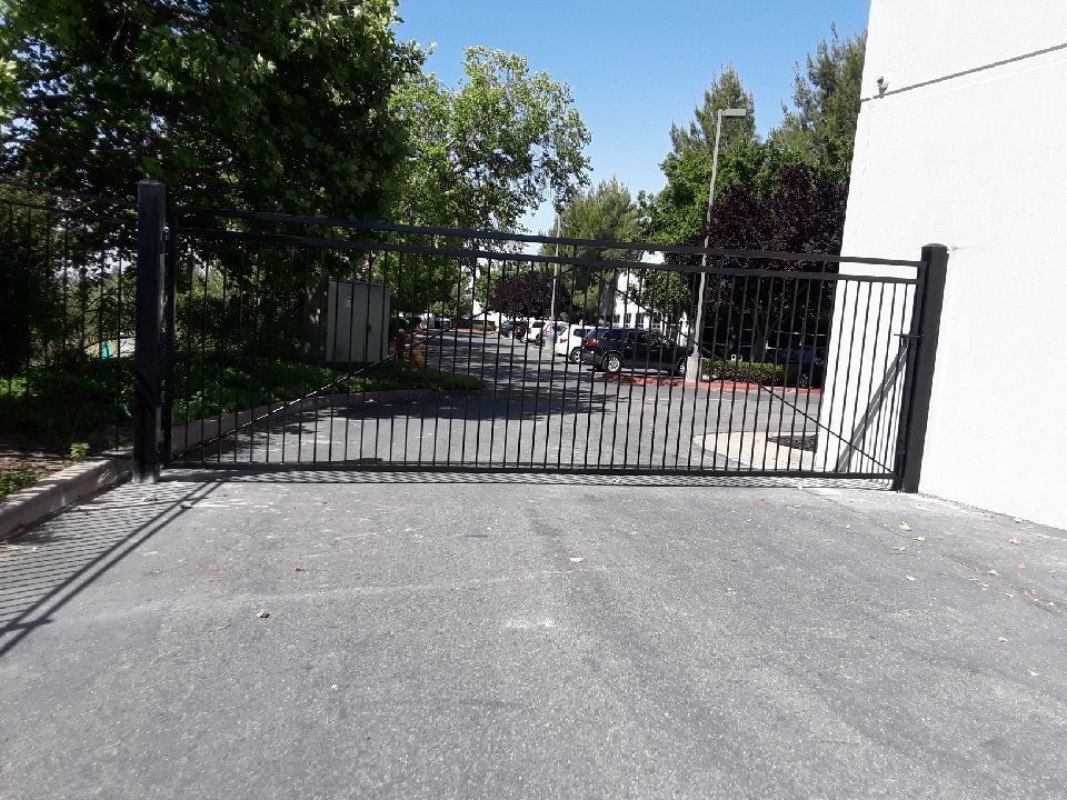 Black metal gate, partly open, blocks a driveway. Asphalt surface. Building and trees in the background under blue sky.