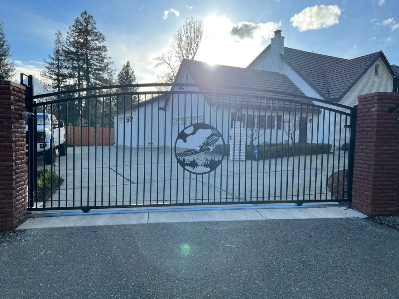 Black metal gate with decorative emblem, brick pillars, and a house in the background.