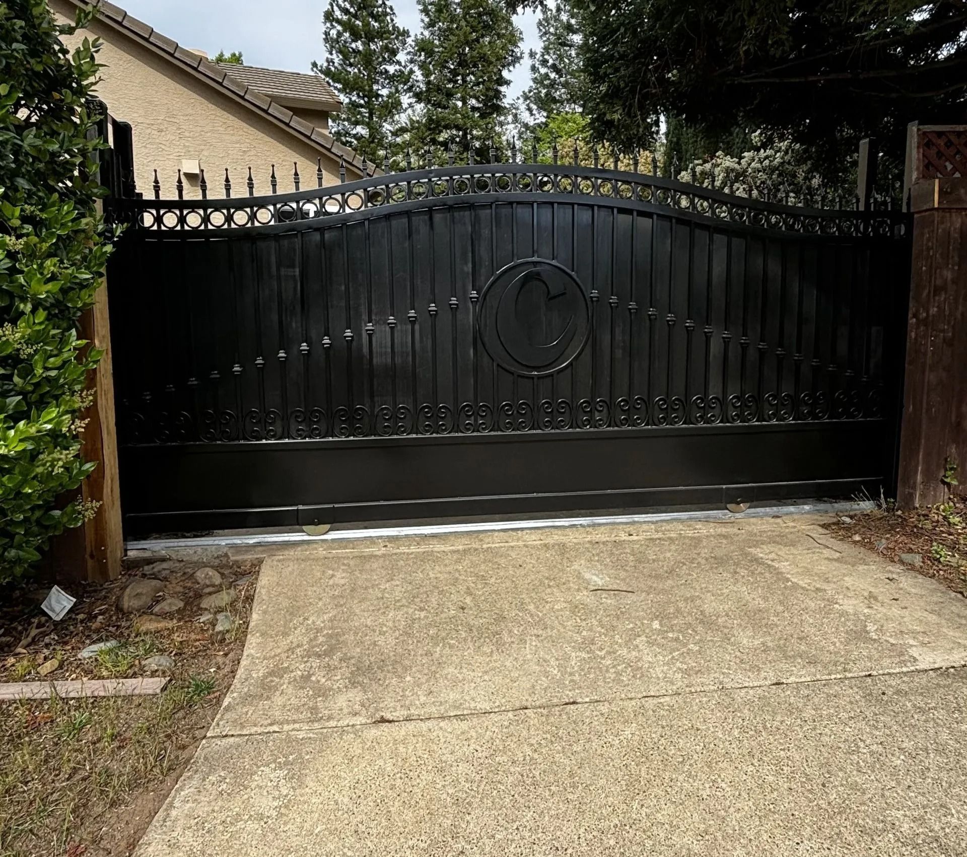 Black metal driveway gate with decorative design and circular emblem. Concrete driveway.
