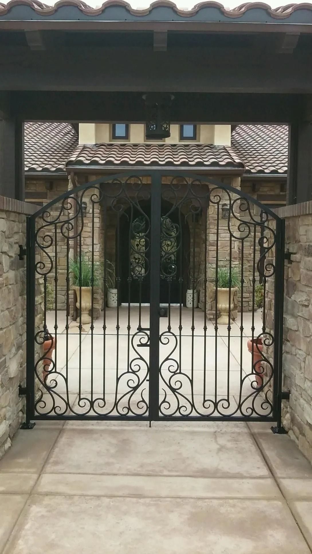 Black wrought iron gate leading to a house with stone walls and a tiled roof.