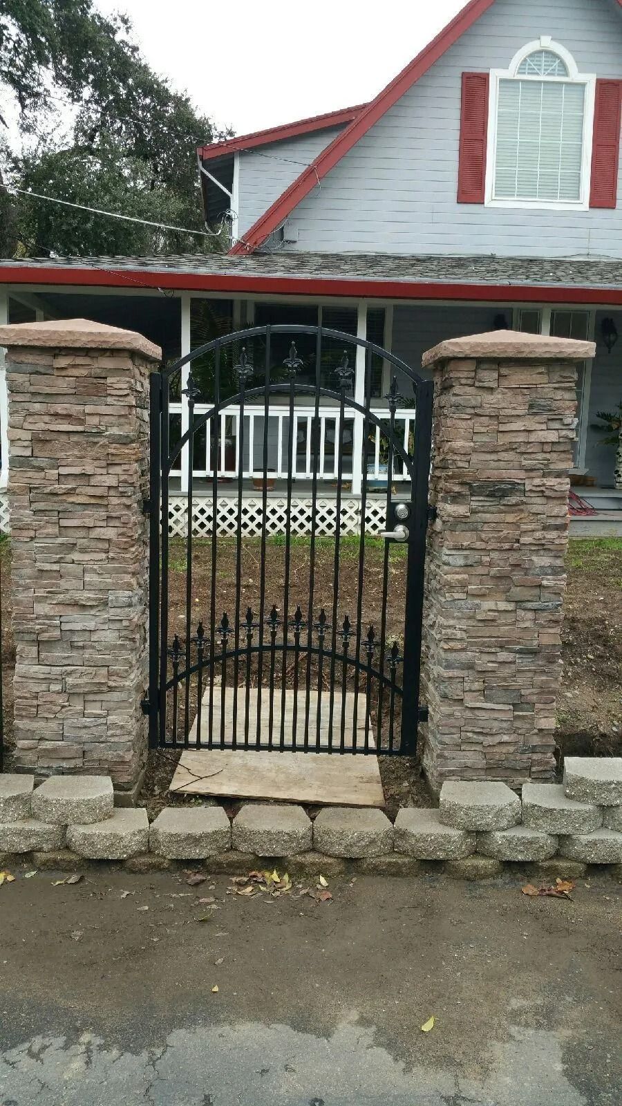 Black iron gate between two stone columns, leading to a gray house with red shutters.