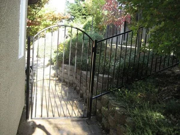 Black metal gate in a landscaped yard, leading to a path alongside a retaining wall.
