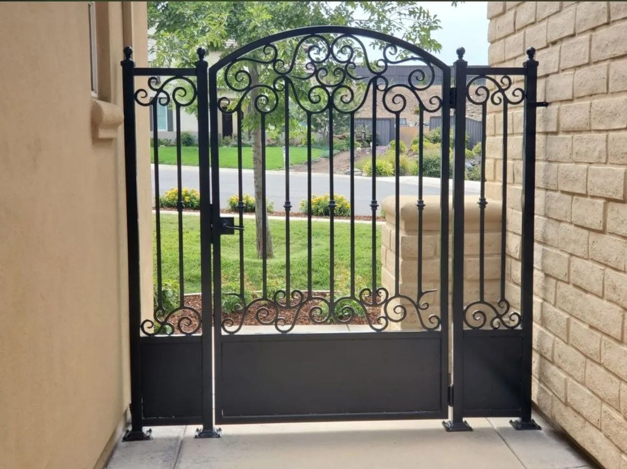 Black wrought iron gate with decorative scrollwork, secured between beige walls on a concrete surface.