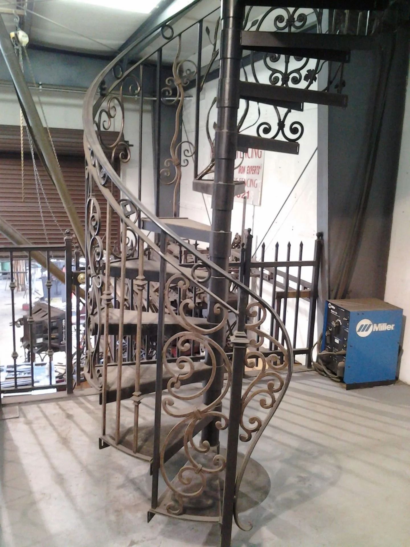 Ornate wrought iron spiral staircase in a workshop setting, with decorative scrollwork and a welding machine nearby.