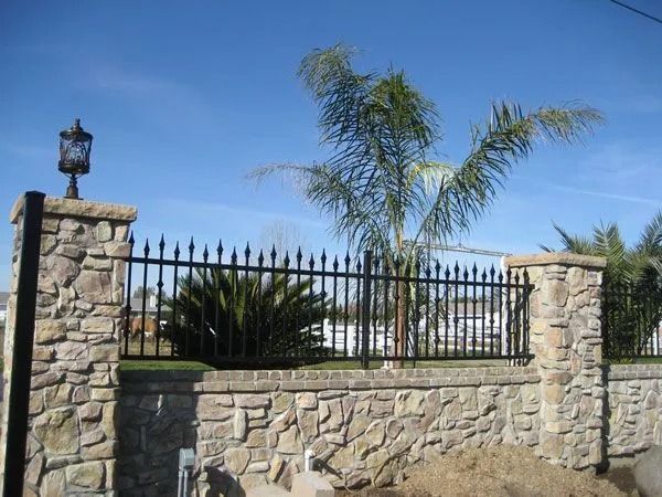 Stone wall with black wrought iron fence, topped with decorative pillars and lamp. Palm tree in background.
