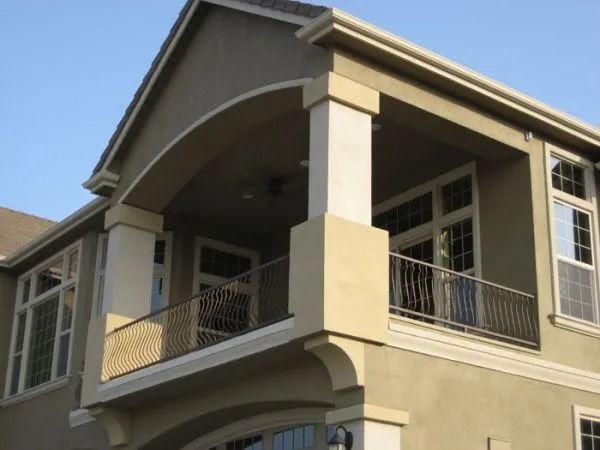 Two-story house with tan stucco exterior, balcony with metal railing, and arched entryway.