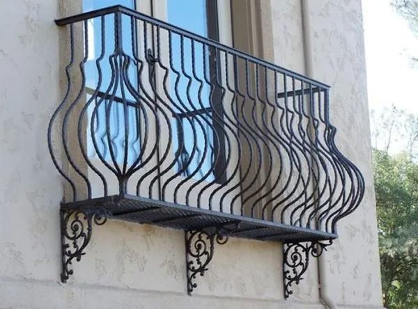 Black wrought iron balcony with decorative curved design against a beige stucco wall, bracket supports below.
