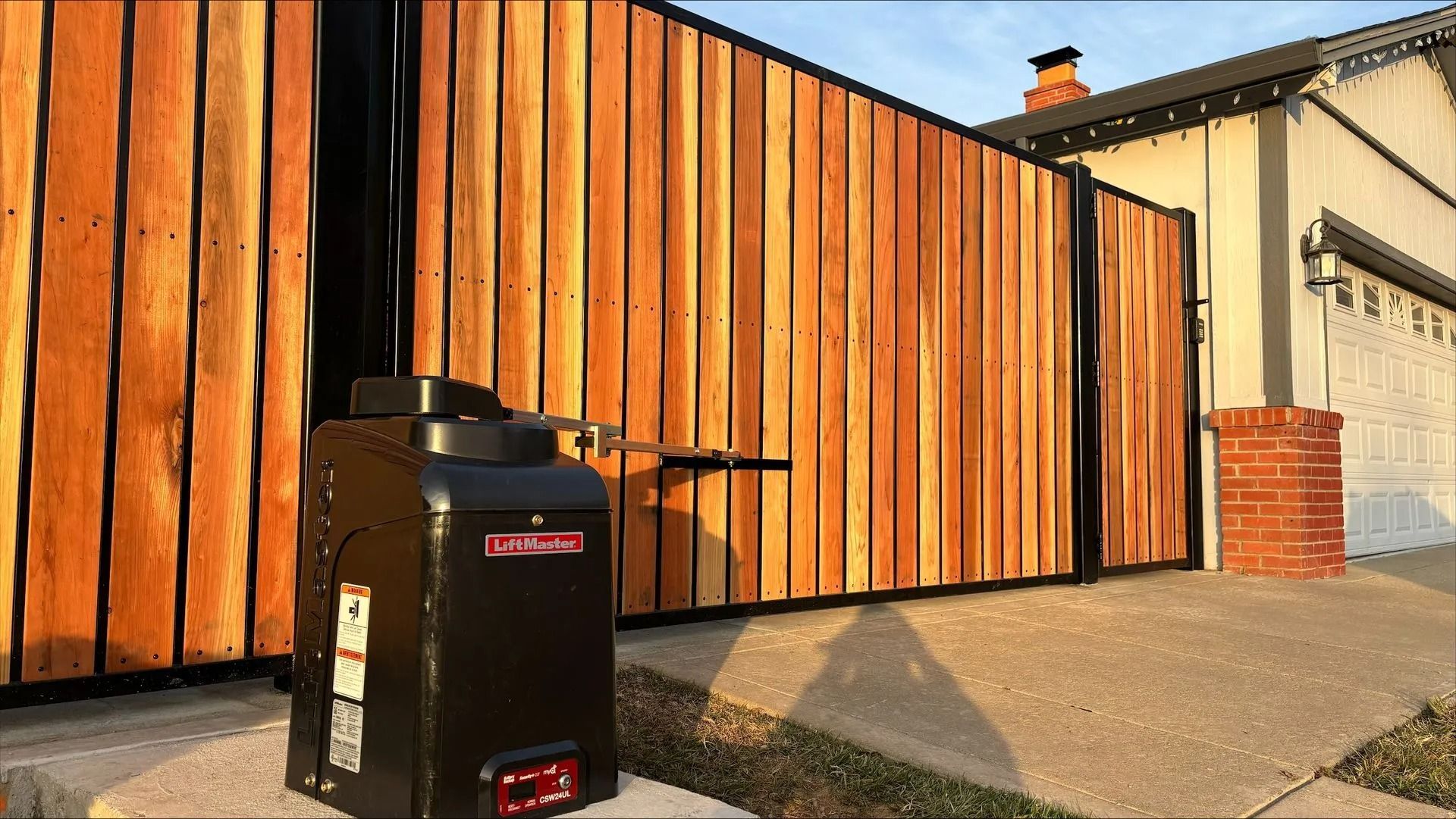 Black gate opener in front of a wooden gate, beside a house with a garage and brick accents.