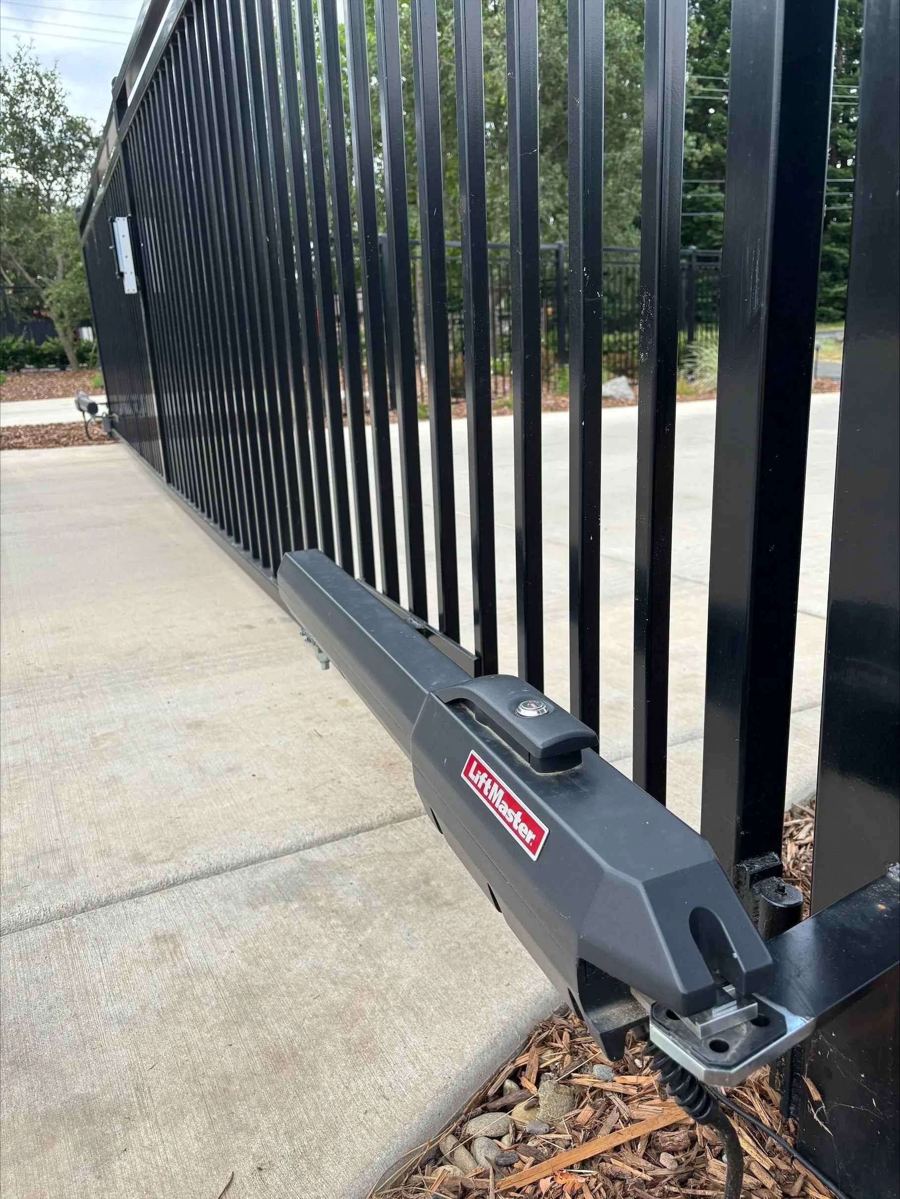 Black automatic gate with an electric opener attached to the front post. Concrete ground.