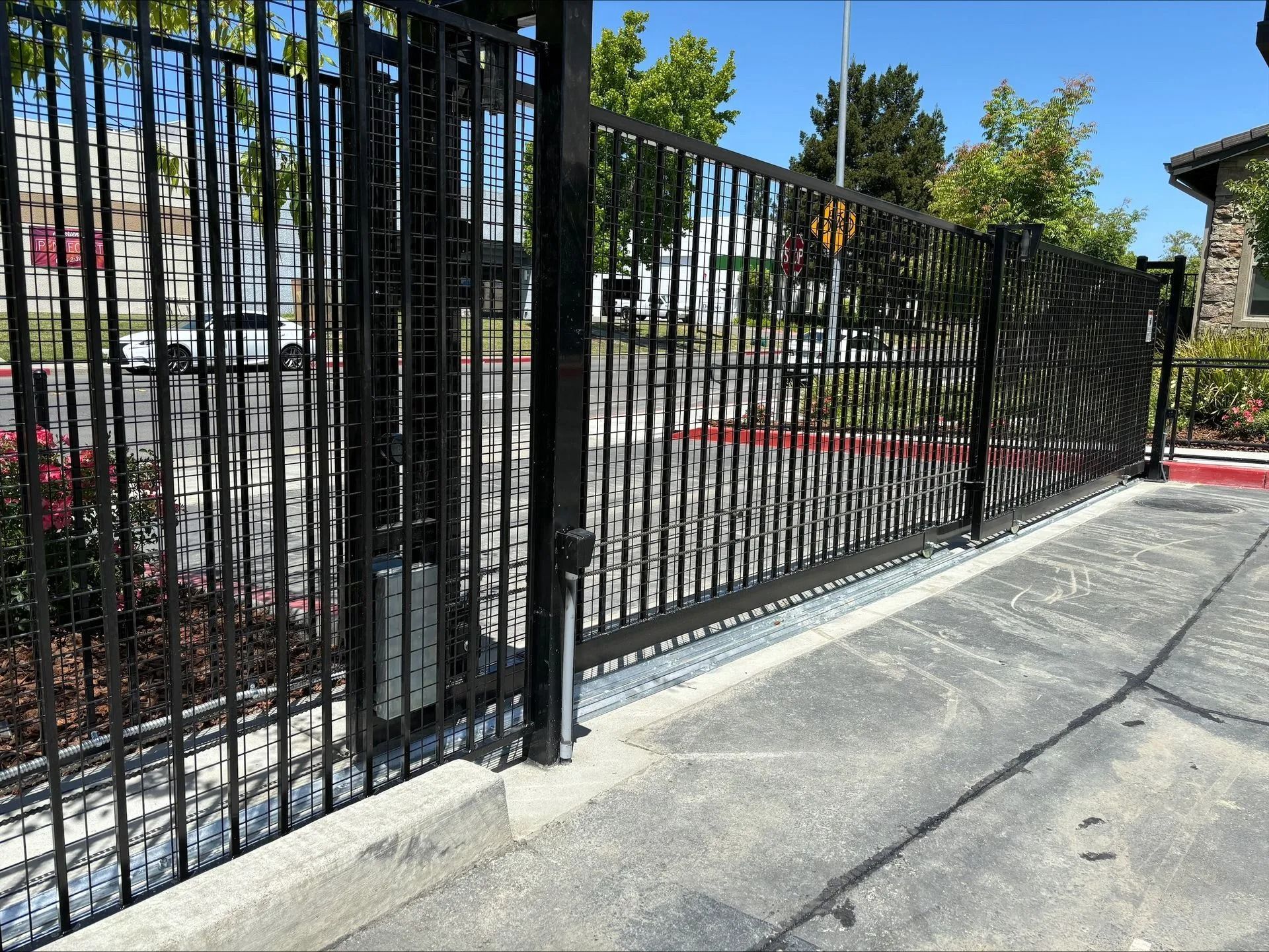 Black metal gate, partly open, spanning a concrete driveway. Sunny day, residential setting.