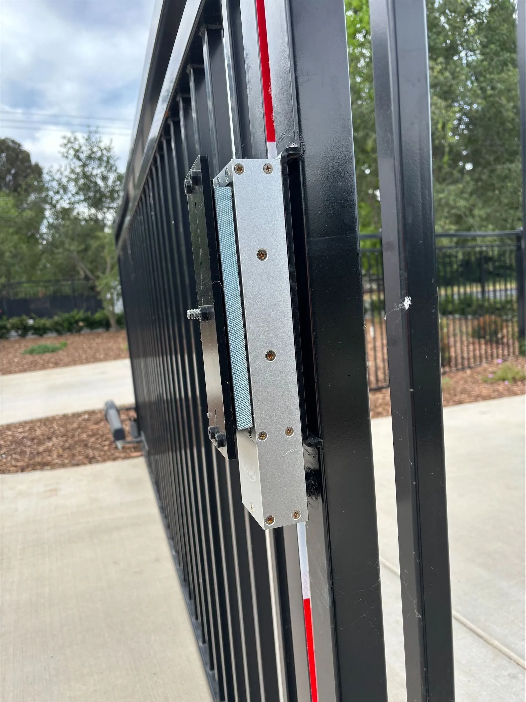 Black metal gate with a silver rectangular locking mechanism, red and white wires, outdoors.