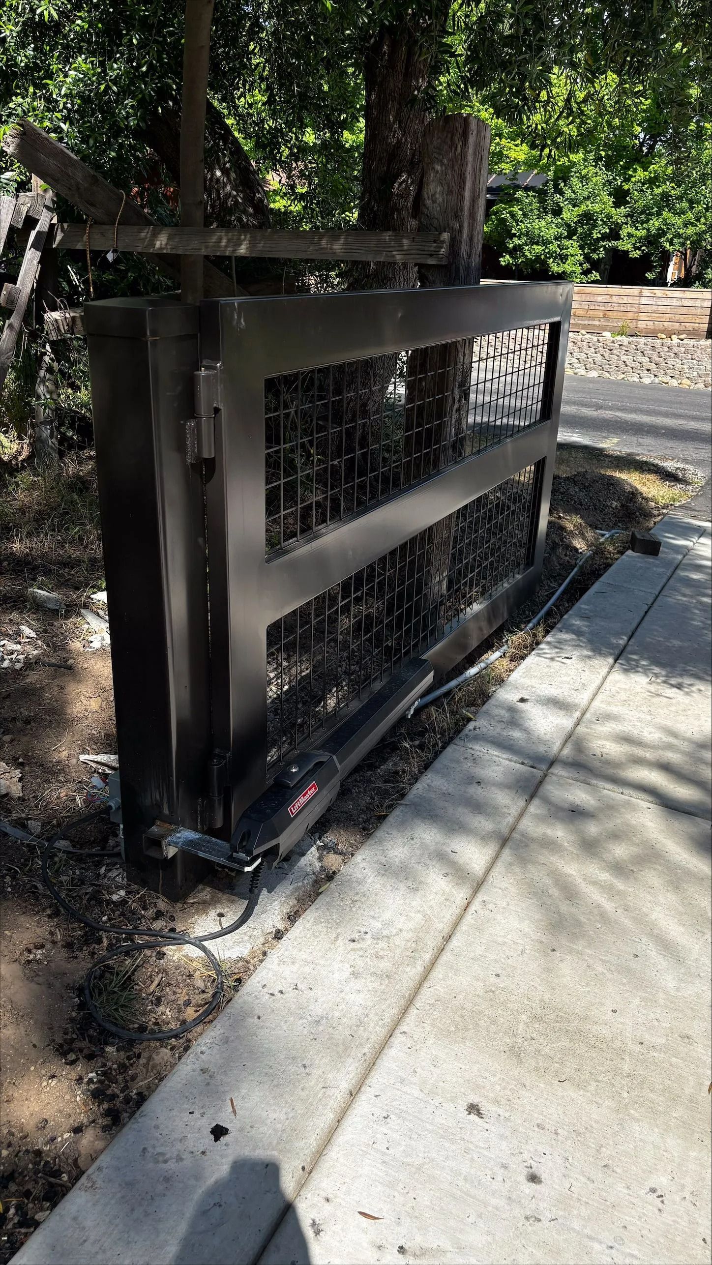 Black metal gate with wire mesh, set on concrete next to a driveway.