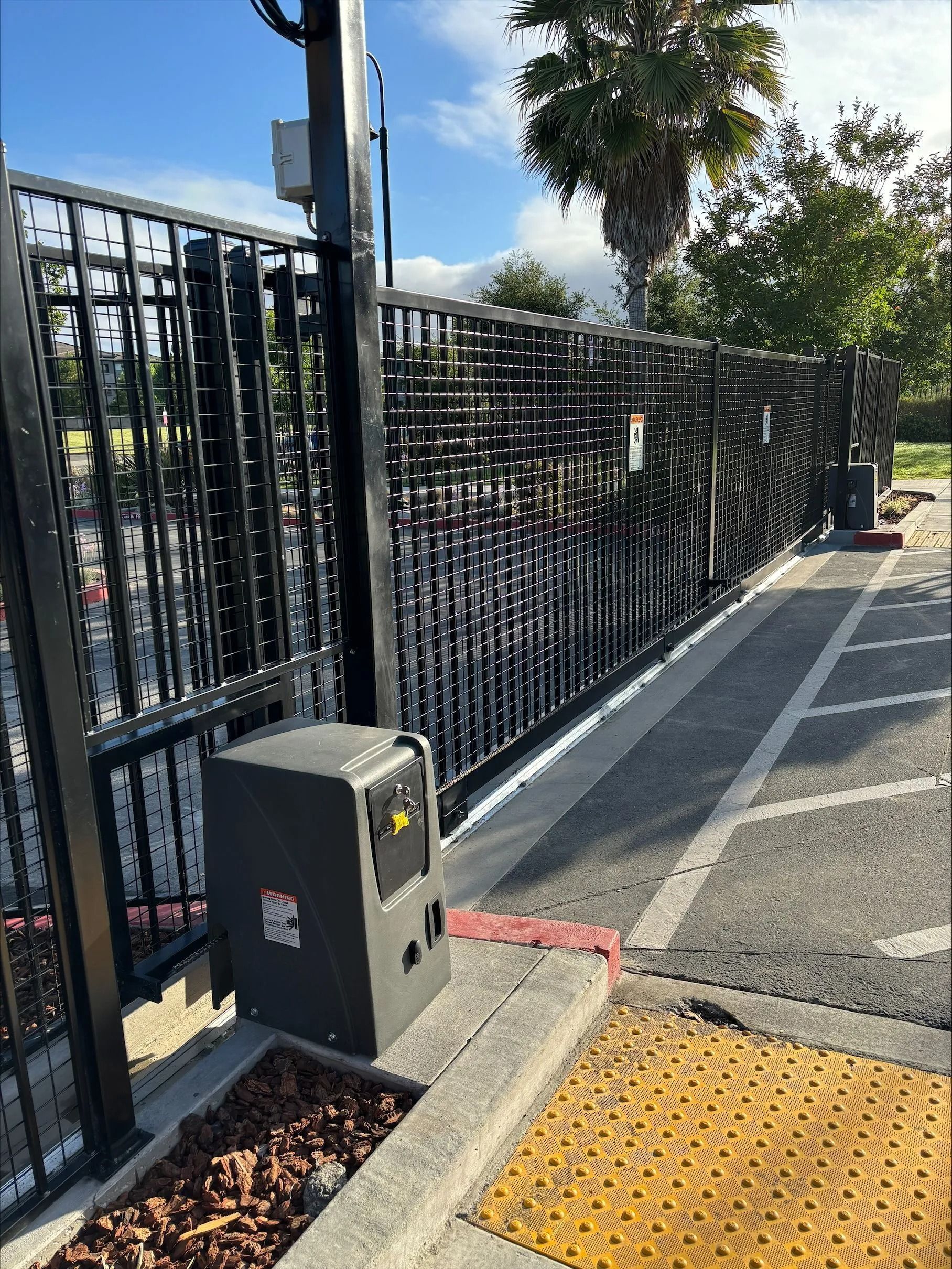 Black security gate with motorized opener on concrete pad next to asphalt.