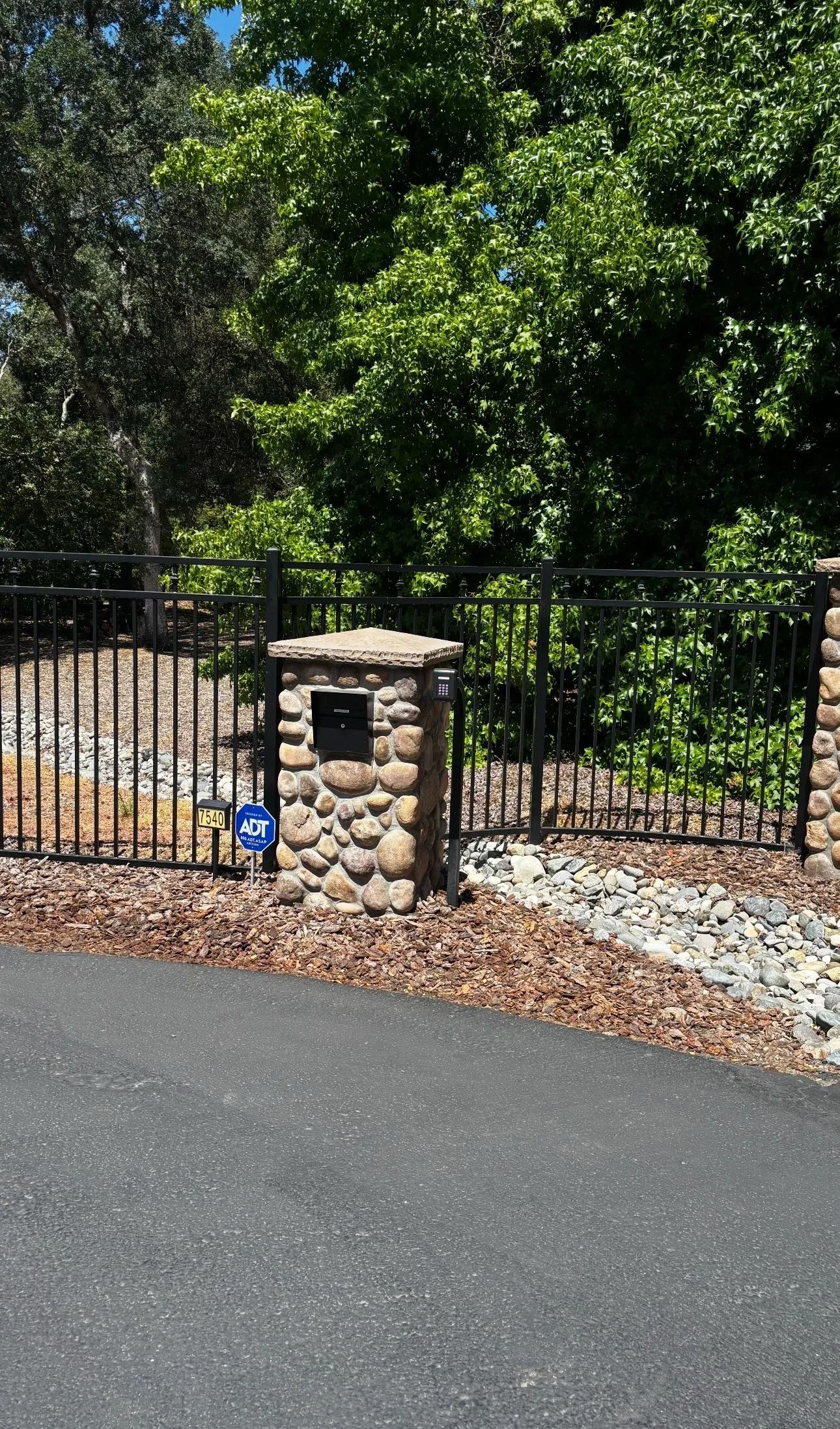 Stone pillars, black wrought iron fence, mailbox, driveway, and trees.