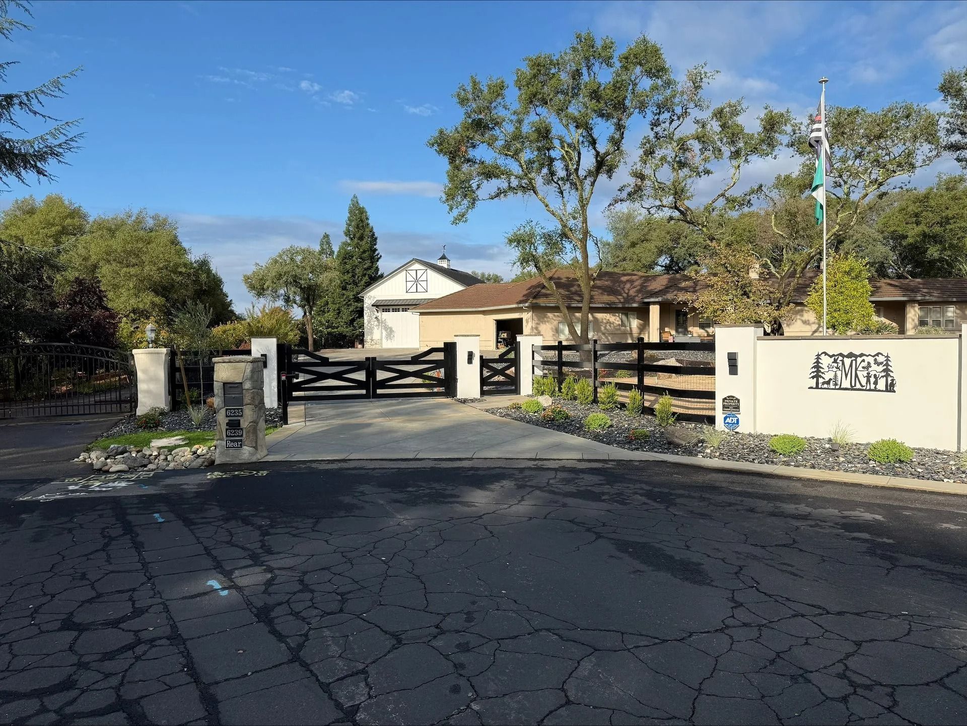 Gated entrance to a ranch-style house on a sunny day. Black gate, white pillars, paved driveway.