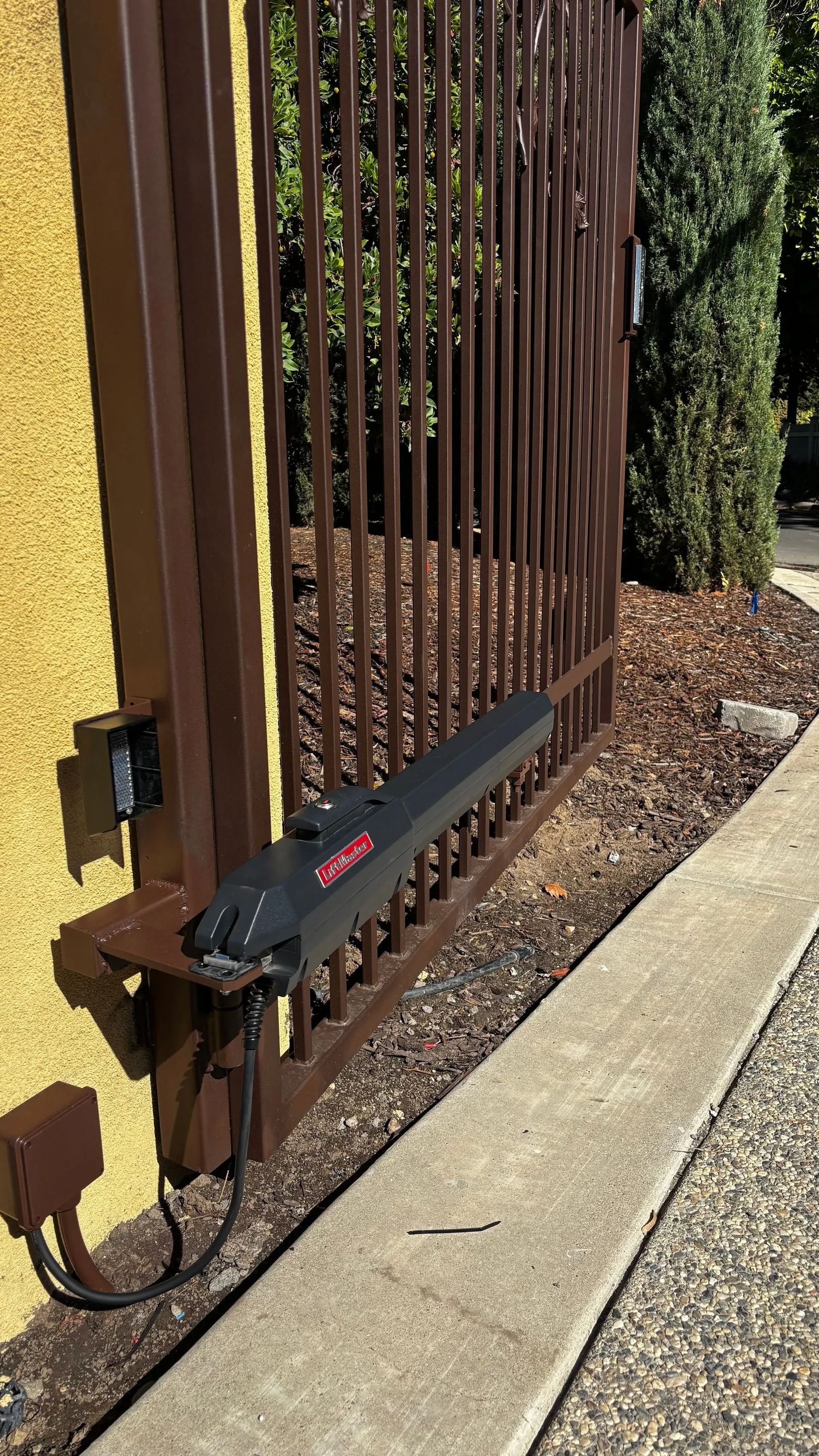 Brown metal gate with automated opener on a concrete driveway.
