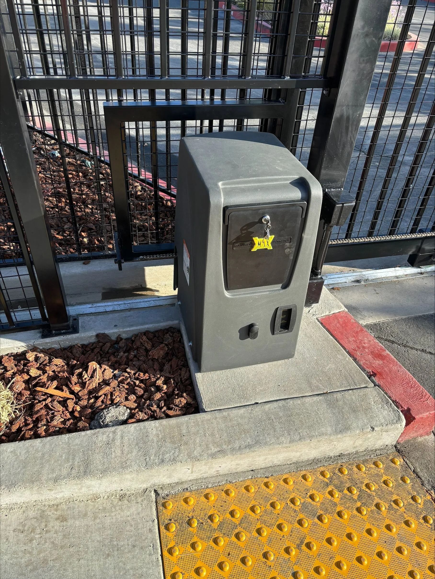 Gray box near a black metal gate, on a concrete surface with yellow tactile paving.