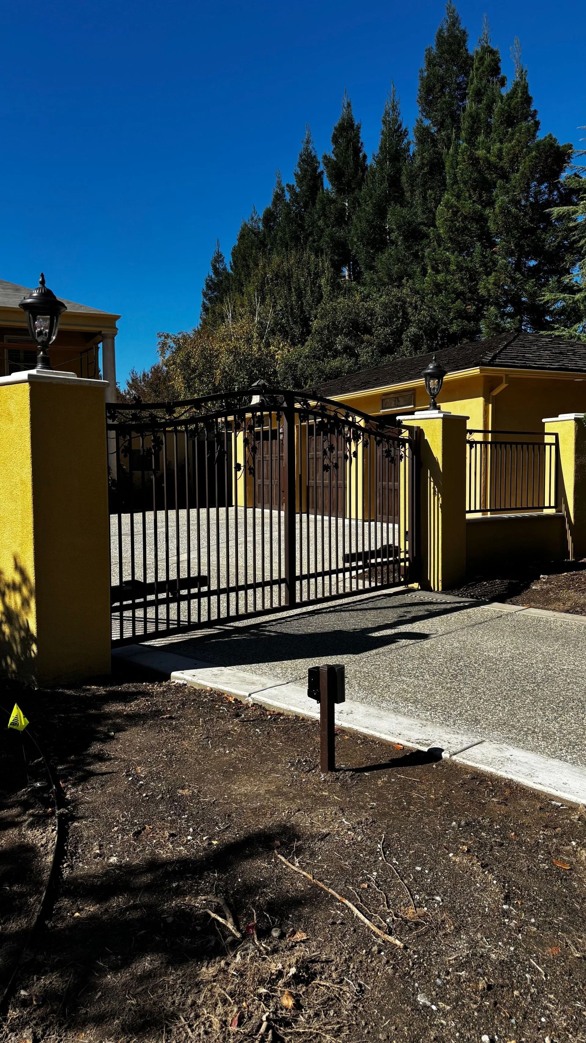 Black metal gate within yellow brick columns, gravel driveway. Trees and blue sky background.