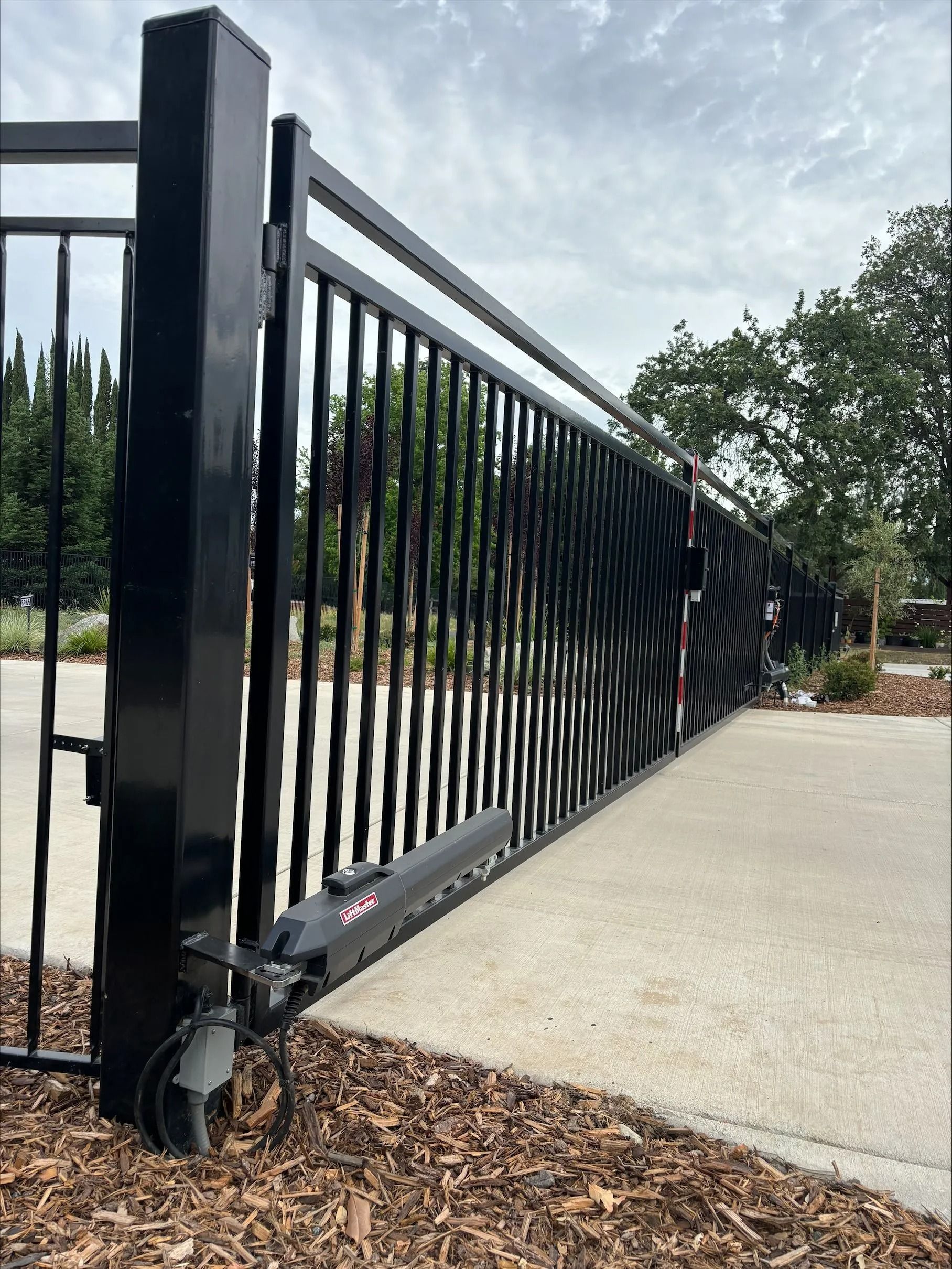 Black metal sliding gate on concrete, opening to a driveway. Vertical bars and horizontal rails.