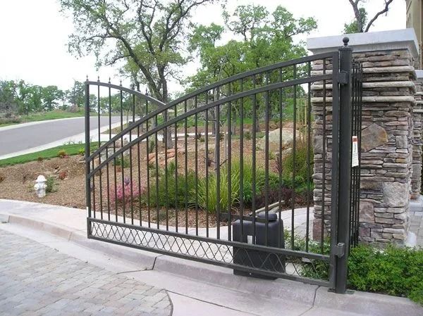 Black metal gate at the entrance to a driveway, opening. Stone pillar on right. Trees and bushes in the background.