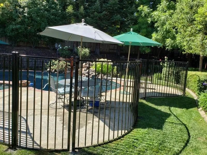 Black fence surrounds a backyard pool with umbrellas, on a sunny day.