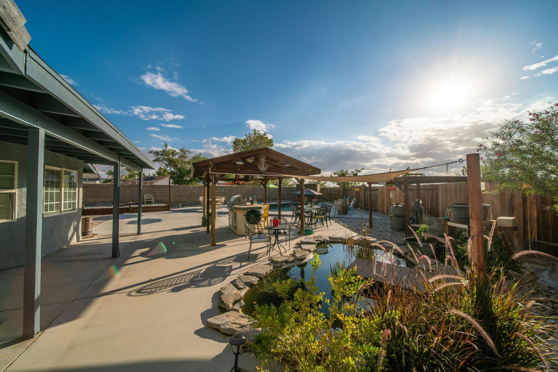 Backyard with pond, wooden pergola, and house under a sunny, partly cloudy sky.