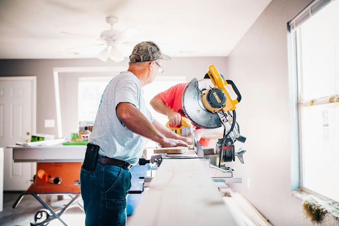 Two people cutting wood with a miter saw in a well-lit room.
