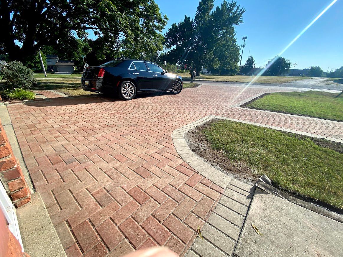 Black car parked on a red brick driveway, with a green lawn and trees under a blue sky.