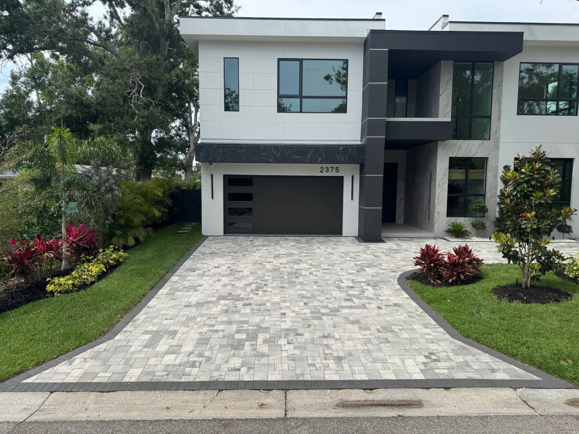 Modern white and gray house with a paver driveway, landscaped yard, and open garage.