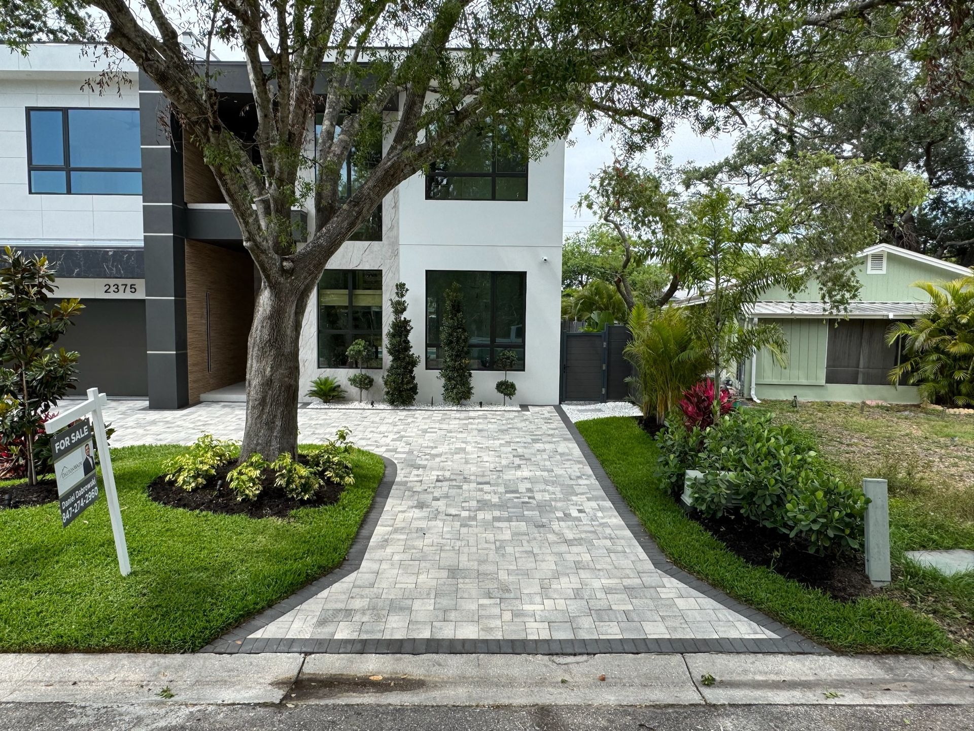 Modern two-story house with paver driveway, lush landscaping, and a for sale sign.