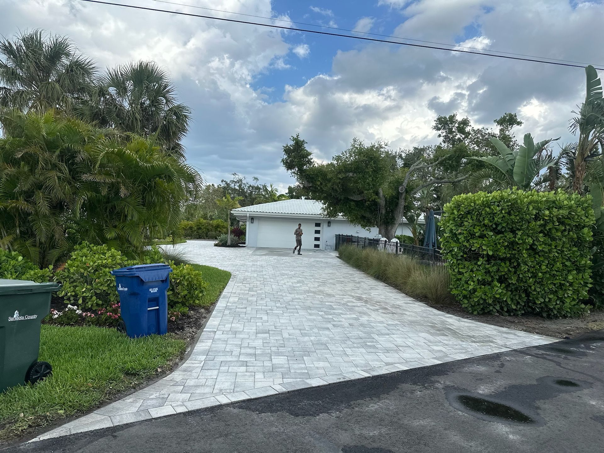 A paved driveway leads to a modern white house with a person standing near the entrance, surrounded by greenery and a blue sky.