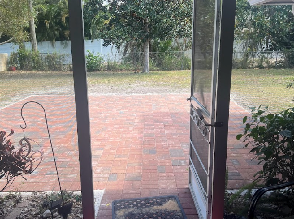 View through an open screen door to a brick patio and backyard with trees and a fence.