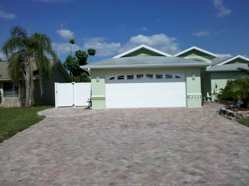 House with white garage door, pale green exterior, and brick driveway under blue sky.